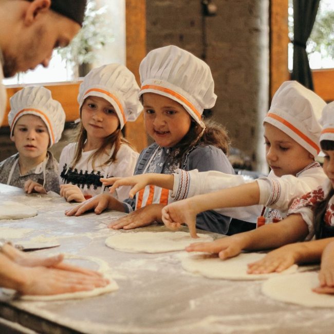 Group of children learning to bake pizza at a restaurant kitchen under a chef's guidance.
